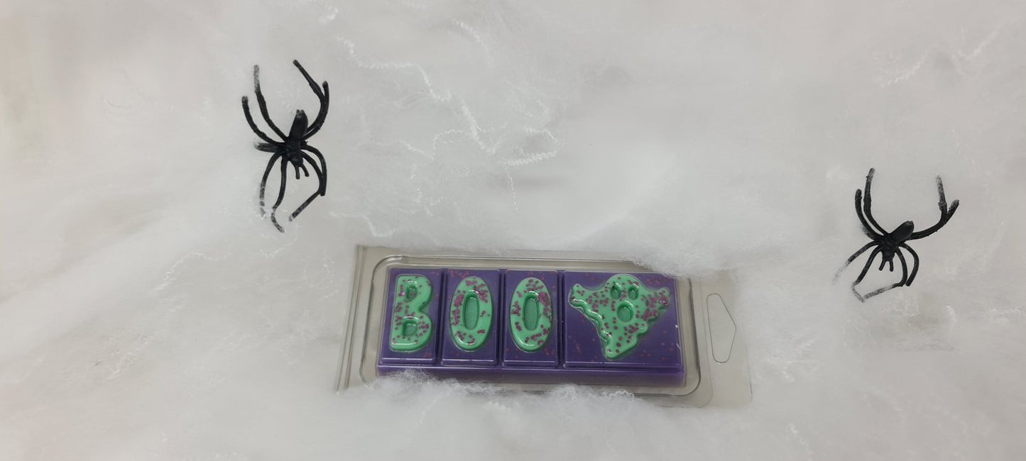 Halloween-themed purple and green wax melt clamshell with "BOO" design surrounded by faux spider webs and plastic spiders.