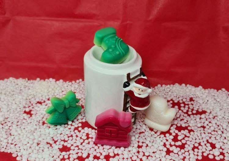 Christmas themed scented wax melts with designs of Santa, bell, stocking, cabin, and snowman, arranged with a white burner on a red background.