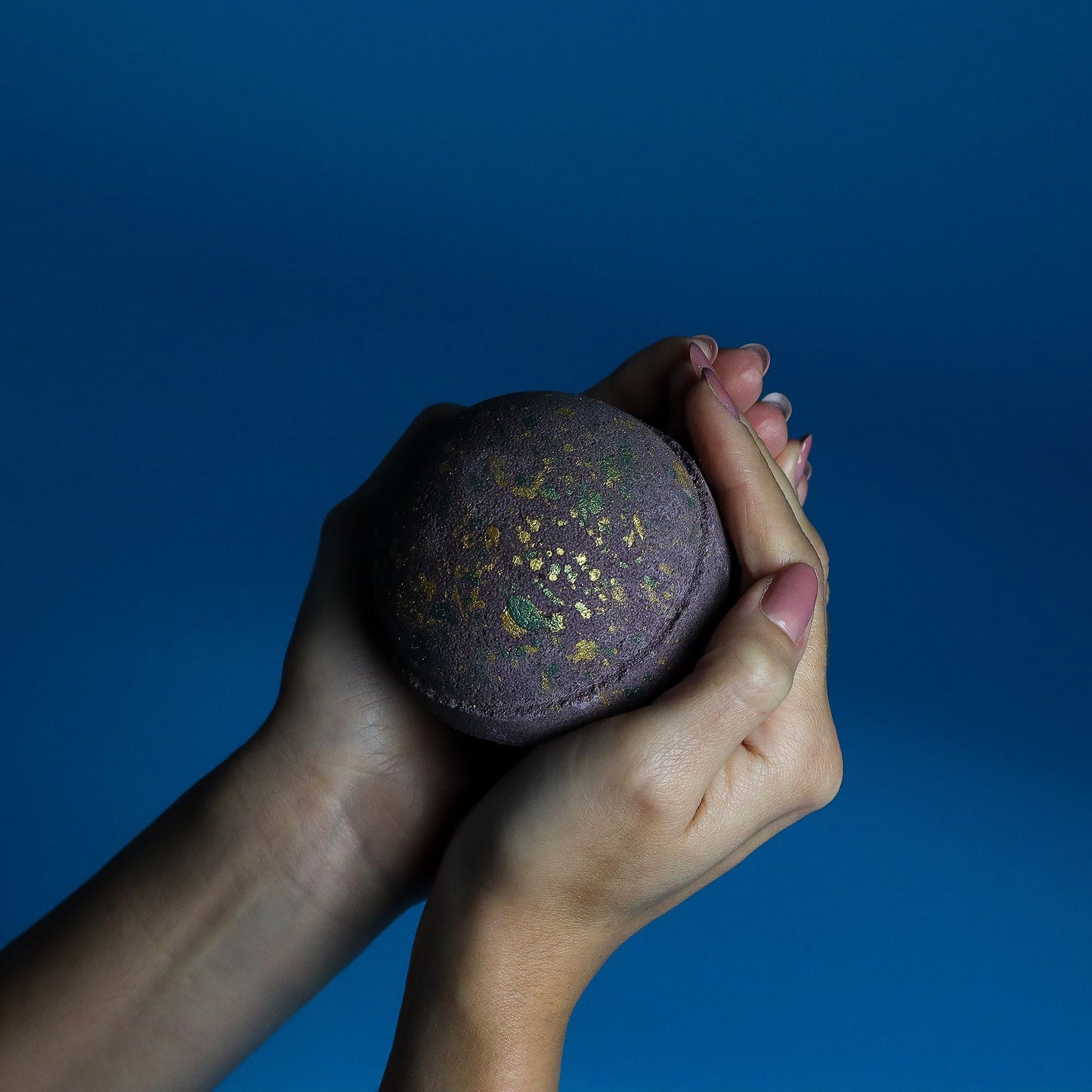 Hands holding Dubai Bath Bomb Emerald Oudh Truffle with gold flecks on dark background