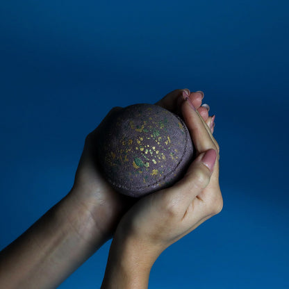 Hands holding Dubai Bath Bomb Emerald Oudh Truffle with gold flecks on dark background