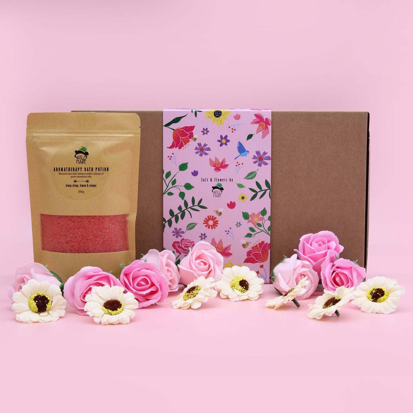 Salt Soak Set with natural pink bath salts and floral packaging surrounded by pink and white flowers on pink background
