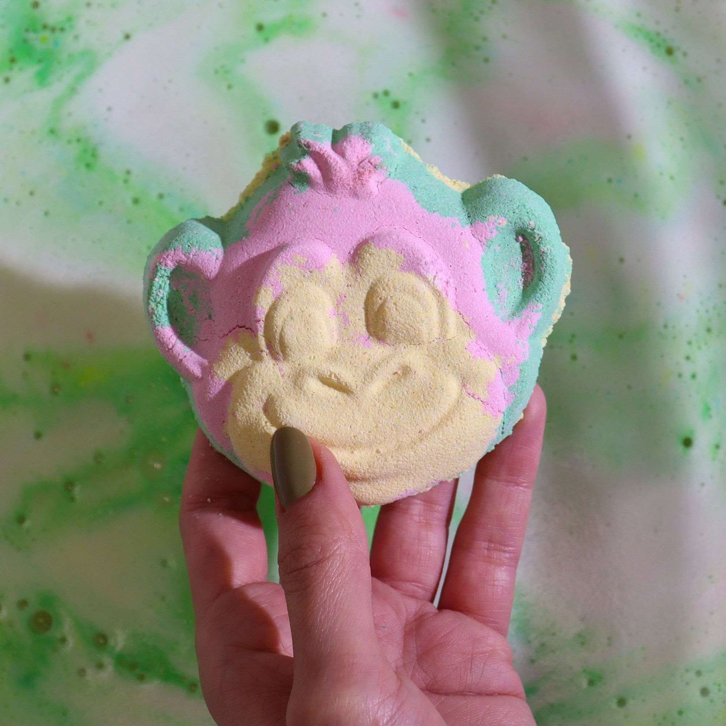 Hand holding colorful monkey bath bomb fizzing in green water