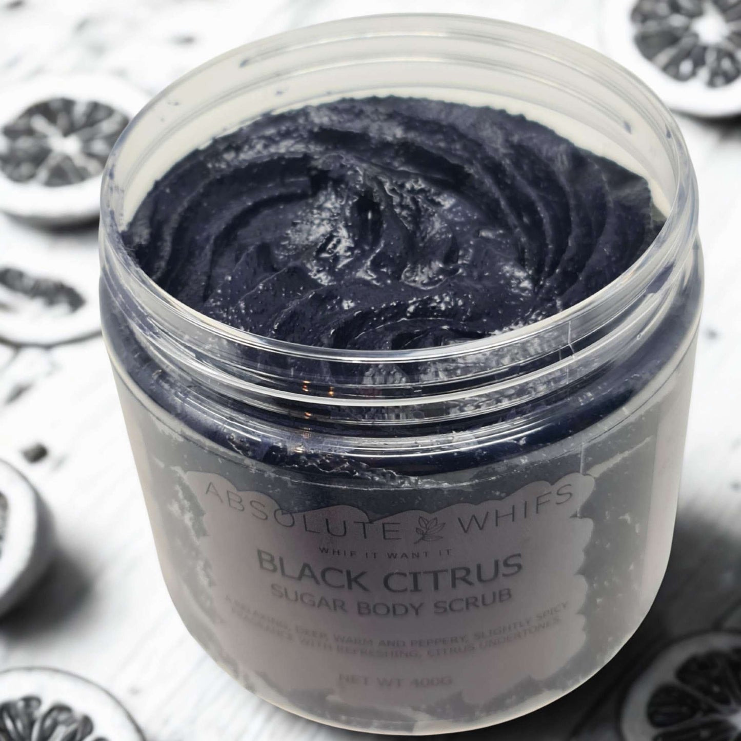 Black Citrus Sugar Body Scrub with dark textured exfoliant in a jar surrounded by black citrus slices