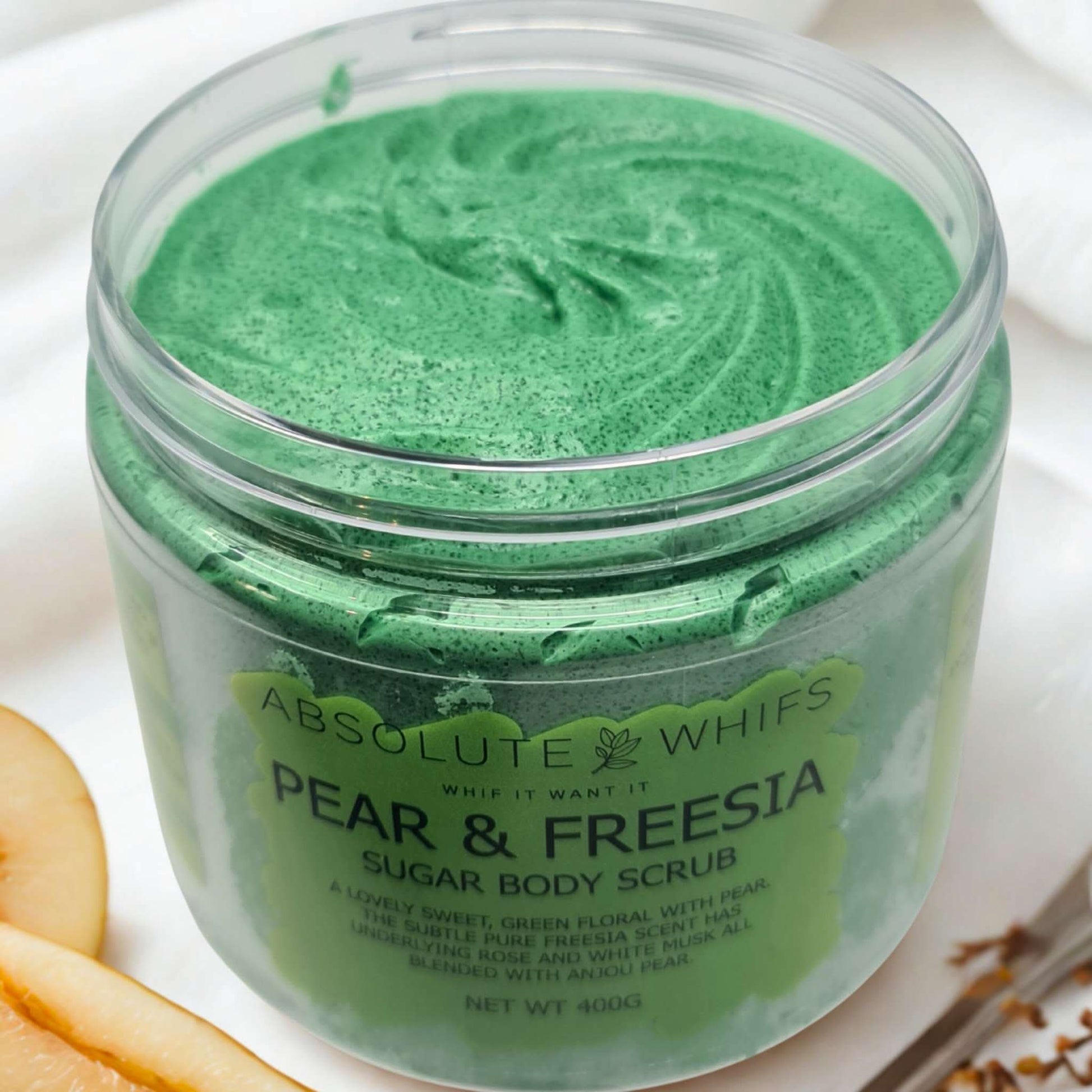 Pear & Freesia Sugar Body Scrub in a clear jar with green scrub and slices of pear nearby