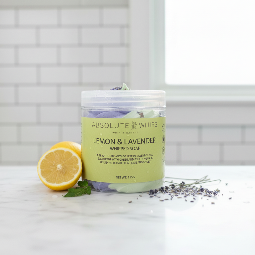 Lemon Lavender Whipped Soap in a small clear jar with light green label, creamy textured soap topped with purple lavender buds and garnished with lemon half and lavender sprigs nearby