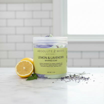 Lemon Lavender Whipped Soap in a small clear jar with light green label, creamy textured soap topped with purple lavender buds and garnished with lemon half and lavender sprigs nearby