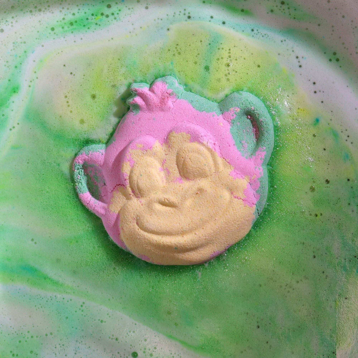 Monkey Bath bomb shaped like a smiling monkey face in pink and beige colors dissolving in green and yellow foamy bath water