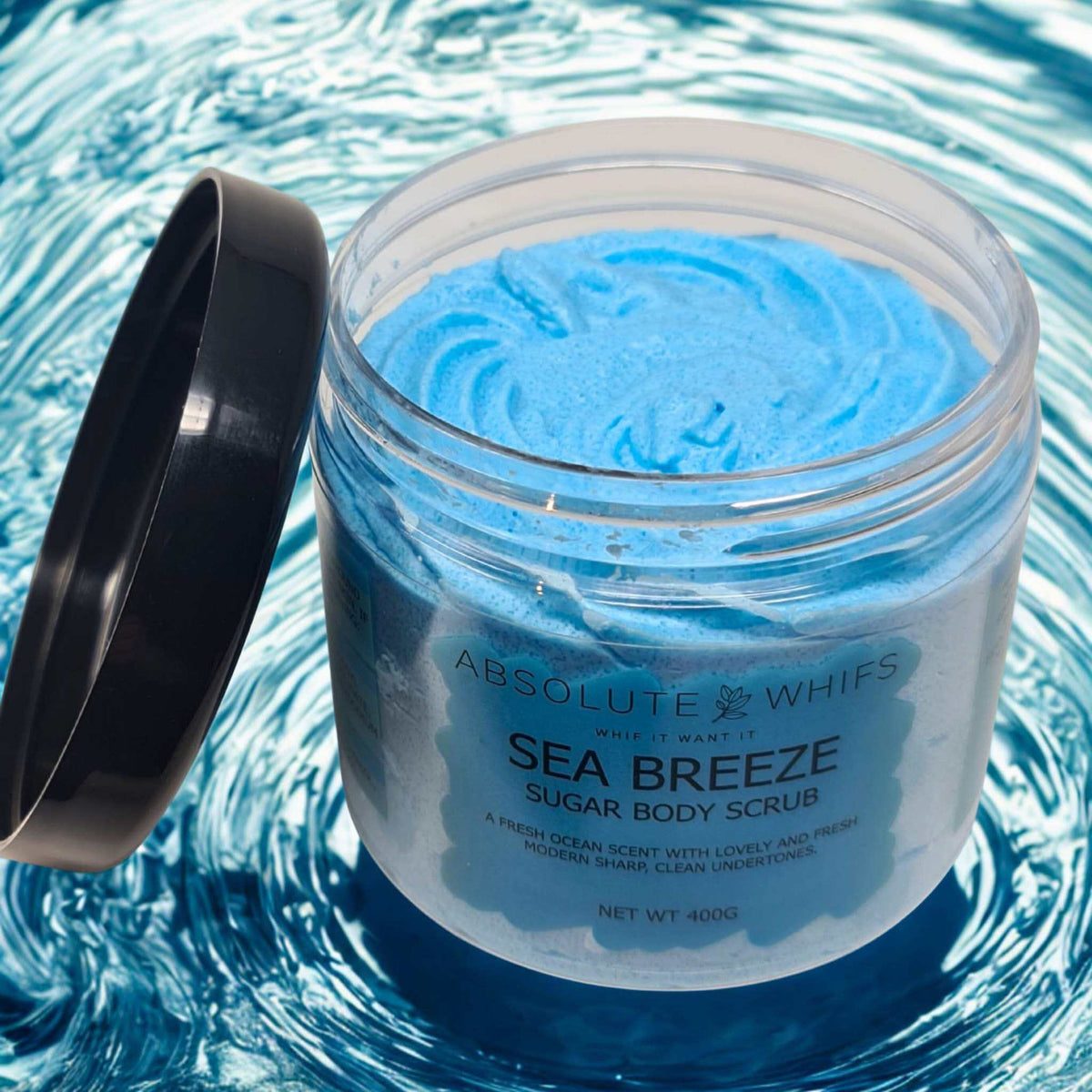 Sea Breeze Sugar Body Scrub in a clear jar with blue whipped texture and black lid on its side over blue water