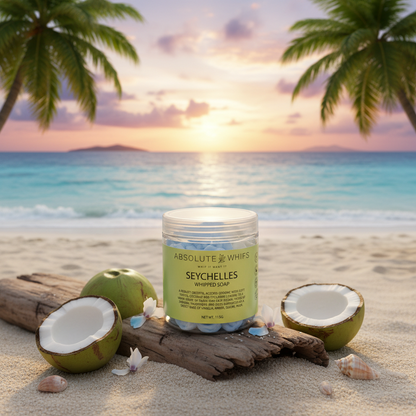 Seychelles Whipped Soap in a clear jar with pale green label placed on driftwood and sand, surrounded by halved coconuts and seashells